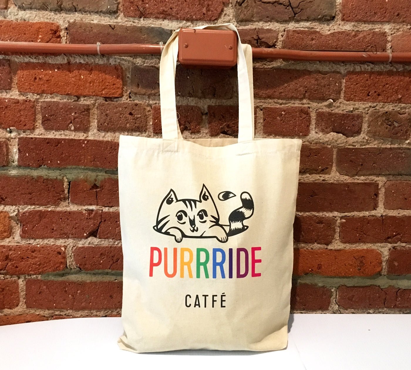 Natural cotton tote bag custom printed with Purrride Catfé cat logo hanging against a red brick wall